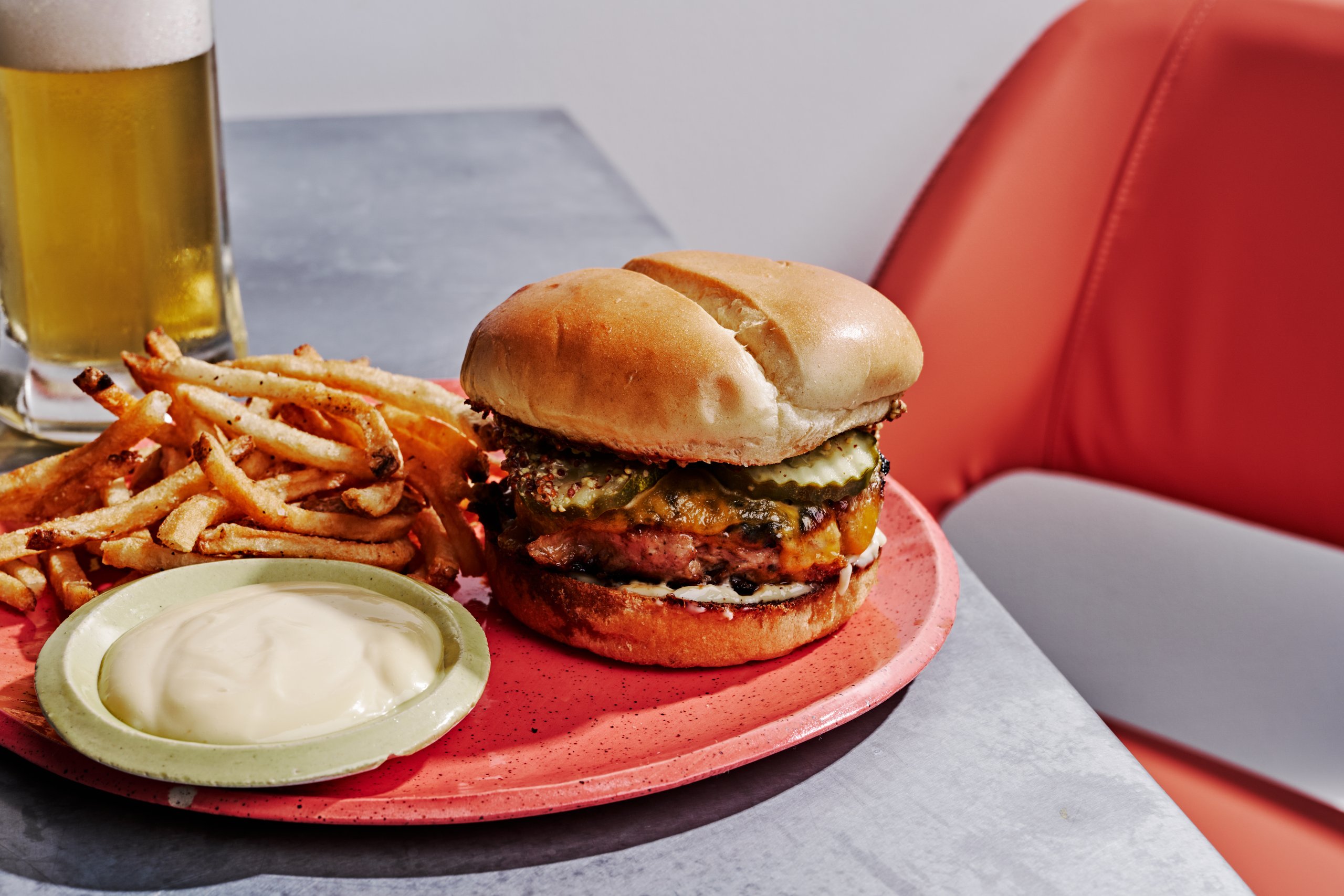 A chicken sandwich with pickles and sauce, served with French fries, mayonnaise, and a glass of beer on a gray table next to an orange chair.