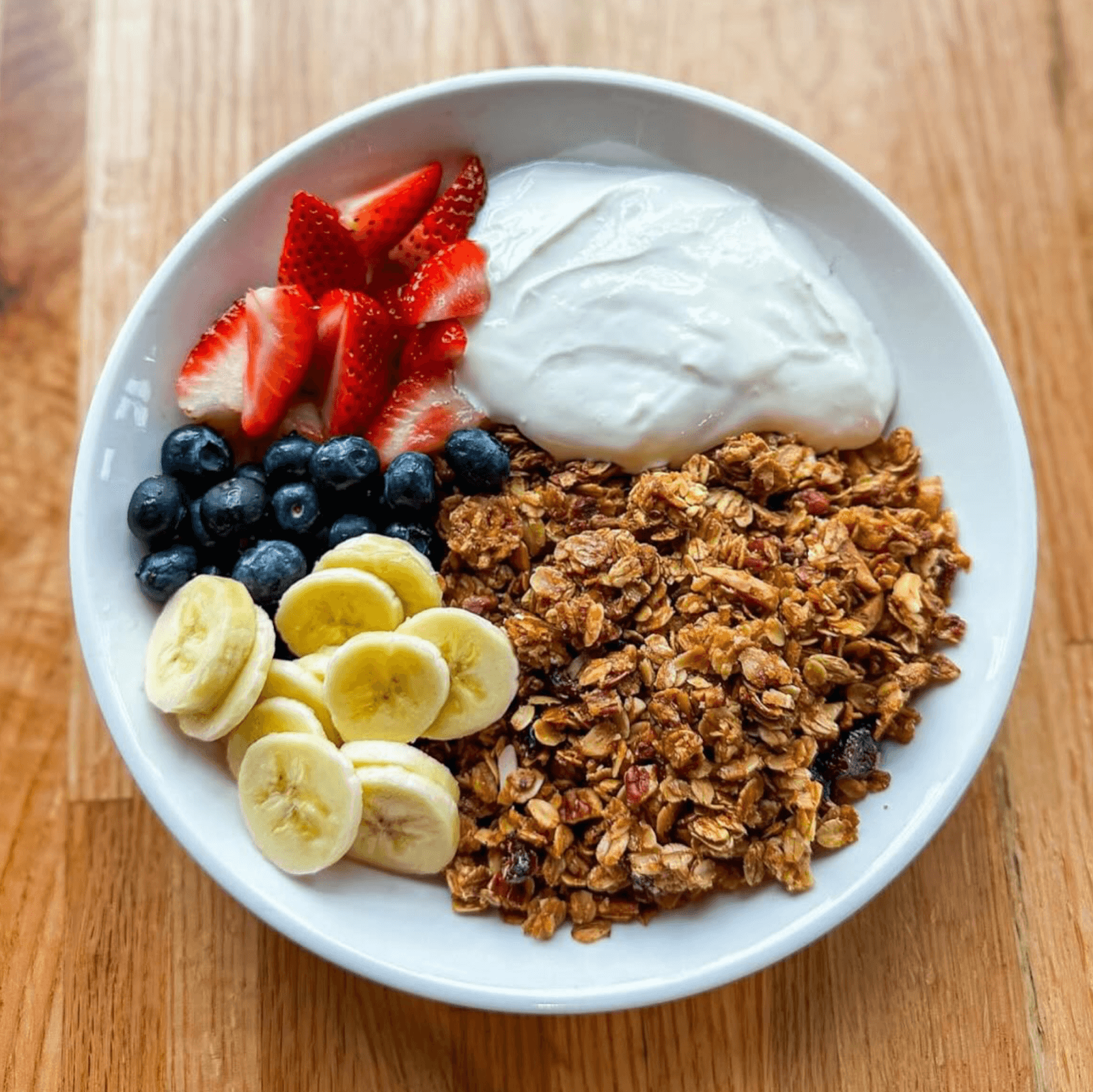 A white bowl containing granola, sliced bananas, blueberries, sliced strawberries, and a serving of plain yogurt on a wooden surface.