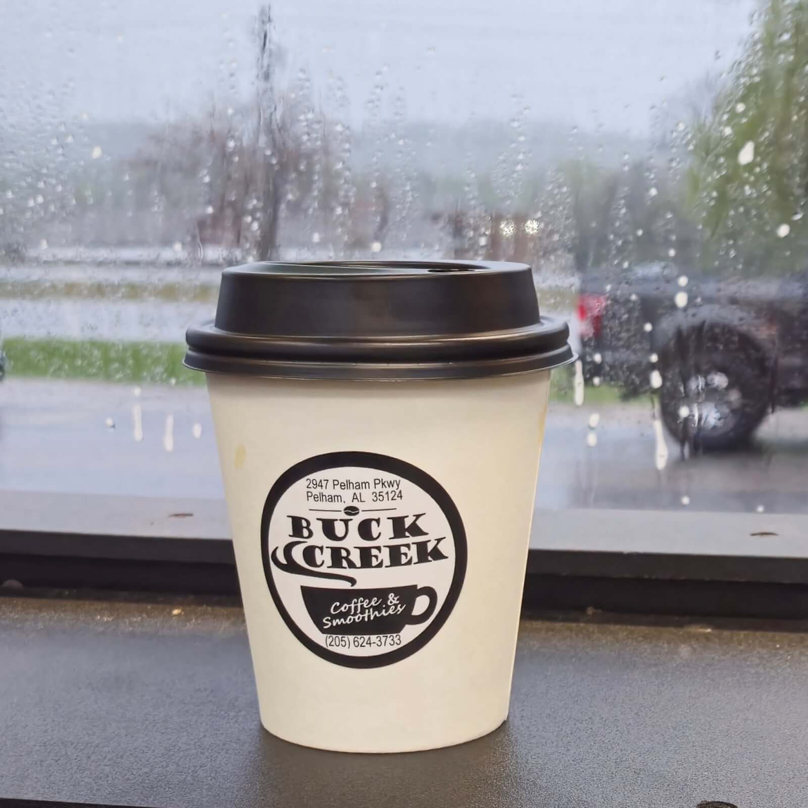 A Buck Creek coffee cup with a lid sits on a windowsill at one of Birmingham's new restaurants; raindrops scatter across the window, framing a blurry outdoor scene in the background.