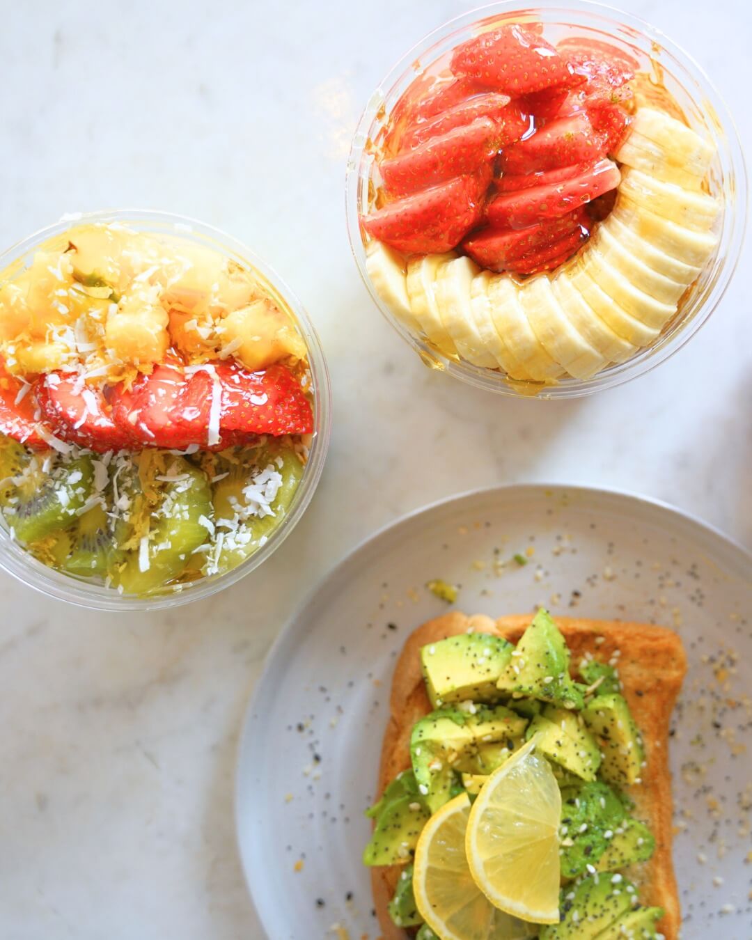 Two fruit smoothie bowls, topped with strawberries, kiwi, banana, pineapple, and coconut, sit beside avocado toast garnished with lemon slices and seeds.
