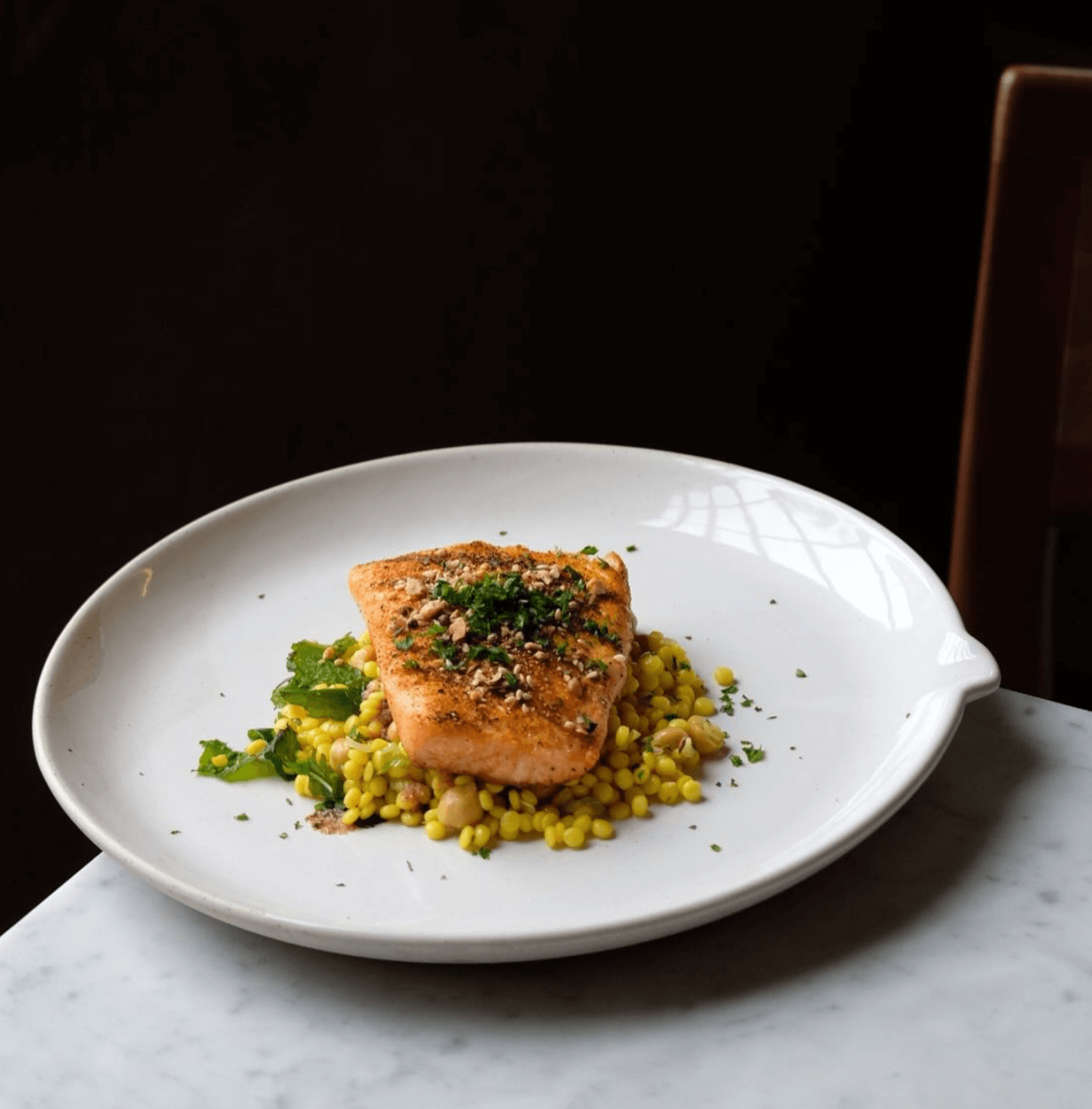 A seared fillet of fish garnished with herbs sits atop pearl couscous and vegetables, beautifully plated in a style inspired by innovative Birmingham restaurants, on a white plate set on a marble table.