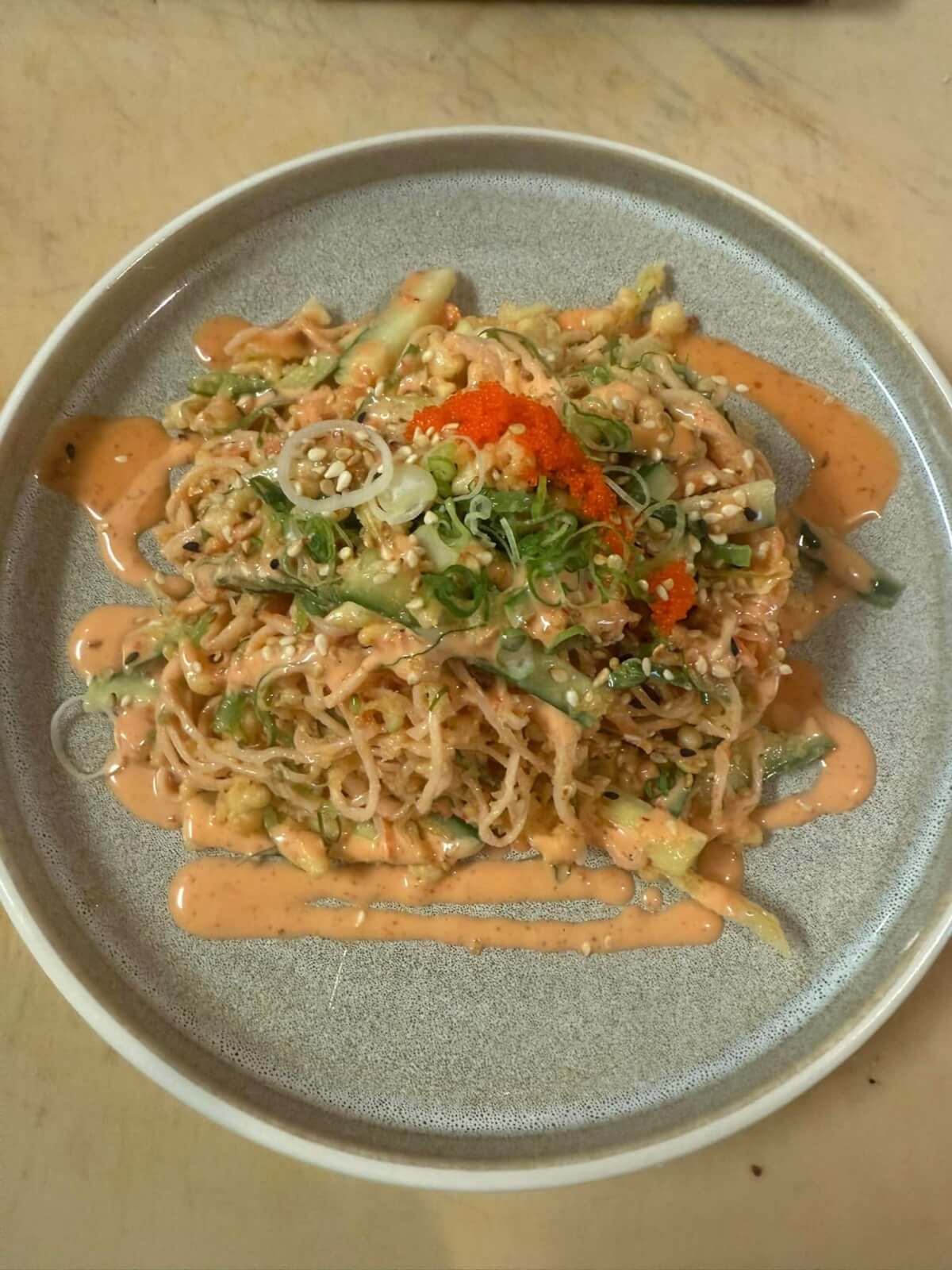 A plate of cold sesame noodles topped with sliced cucumber, green onions, sesame seeds, orange roe, and creamy sauce drizzle.