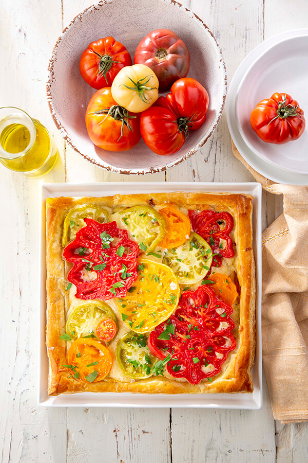 A square tomato tart with sliced heirloom tomatoes on puff pastry sits on a white table next to a bowl of whole tomatoes, olive oil, and plates.