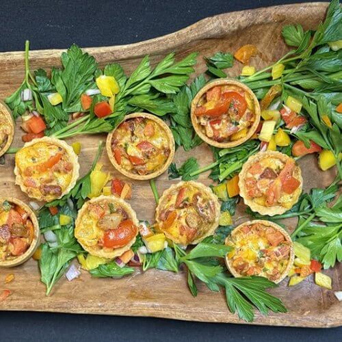 A wooden tray with mini quiches garnished with chopped red and yellow bell peppers, onion, and parsley on a black background.