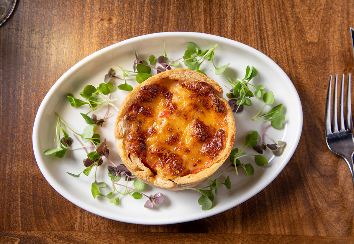 A small baked quiche is served on an oval white plate with fresh microgreens, on a wooden table next to a fork and knife.