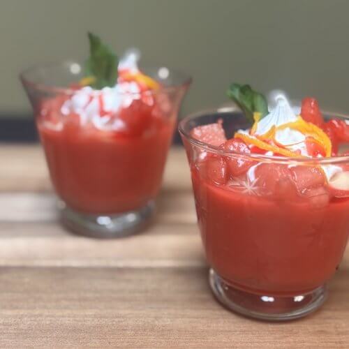 Two glasses filled with a red dessert topped with whipped cream, orange peel, and a green garnish, placed on a wooden surface.
