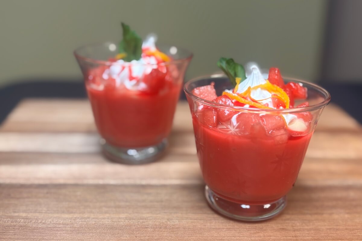 Two glasses filled with a red dessert topped with whipped cream, orange peel, and a green garnish, placed on a wooden surface.