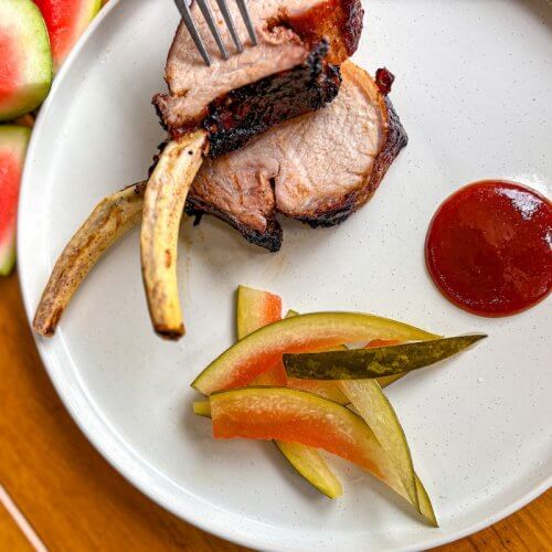 A plate with two pork rib chops, pickled vegetables, a serving of barbecue sauce, and watermelon slices on the side.
