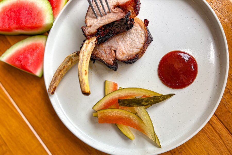 A plate with two pork rib chops, pickled vegetables, a serving of barbecue sauce, and watermelon slices on the side.