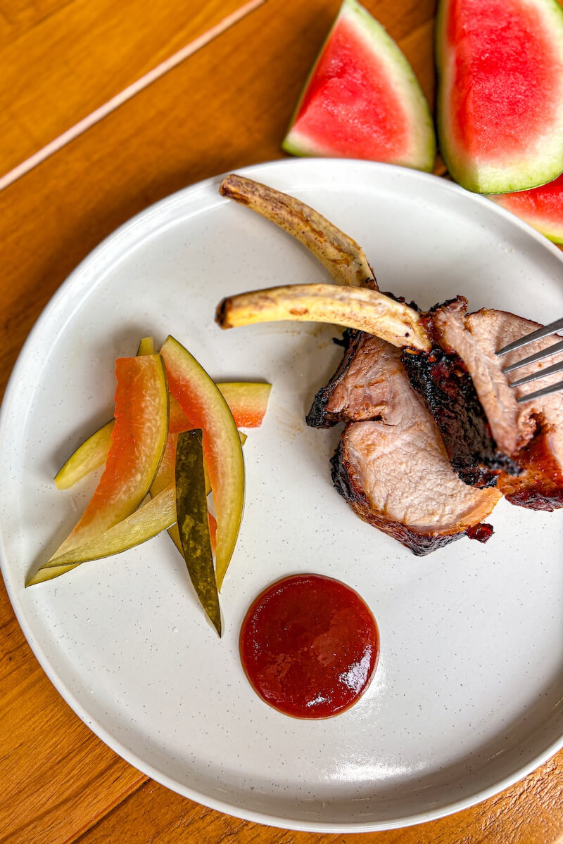 A plate with grilled pork chops, barbecue sauce, sliced pickles, and pickle spears. Watermelon slices are visible in the background.