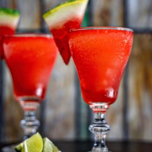 Two glasses of bright red frozen drinks garnished with watermelon slices, with lime wedges placed on the table in front.