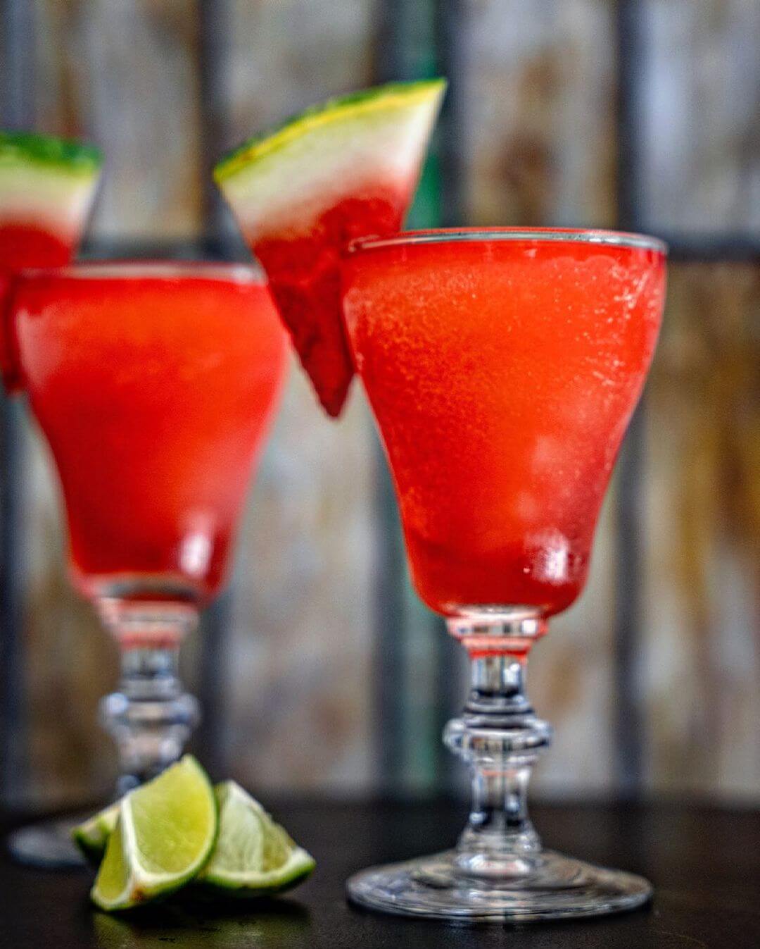Two glasses of bright red frozen drinks garnished with watermelon slices, with lime wedges placed on the table in front.