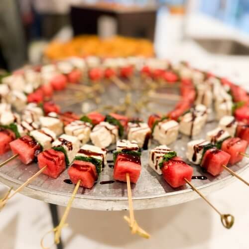 Skewers with cubes of watermelon, mozzarella, and basil, drizzled with balsamic glaze, arranged in a circular pattern on a metal tray.