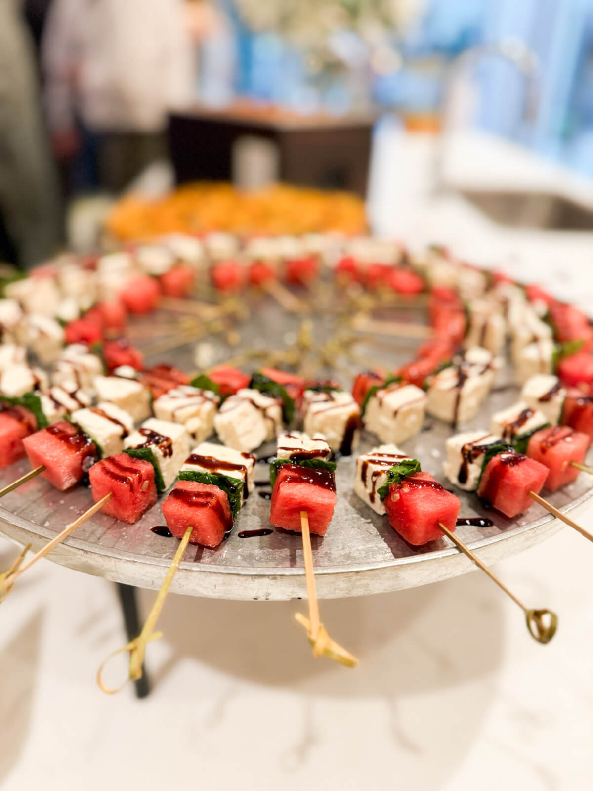 Skewers with cubes of watermelon, mozzarella, and basil, drizzled with balsamic glaze, arranged in a circular pattern on a metal tray.