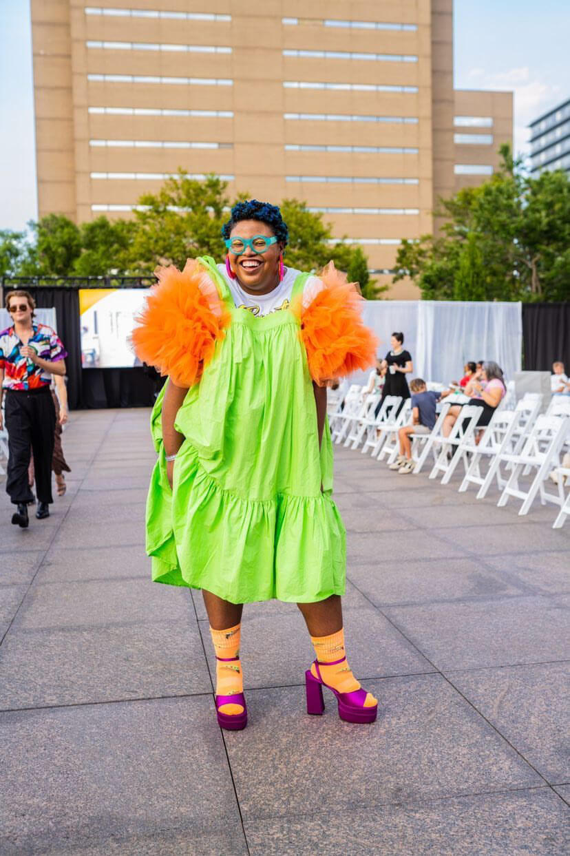 She's Behind BHAM's Most Colorful (and Secretly Nostalgic) Jewelry Line - 1 Aaliyah Taylor in a bright green dress with orange ruffled sleeves, colorful accessories and platform shoes stands smiling outdoors surrounded by white chairs.