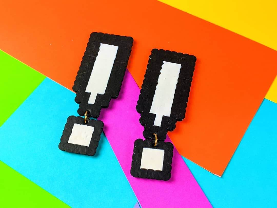 She's Behind BHAM's Most Colorful (and Secretly Nostalgic) Jewelry Line - 6 A pair of pixel art-style exclamation mark earrings in black and white, displayed on a colorful, layered paper background. Exalting in Beauty, these are a striking creation by jewelry designer Aaliyah Taylor.