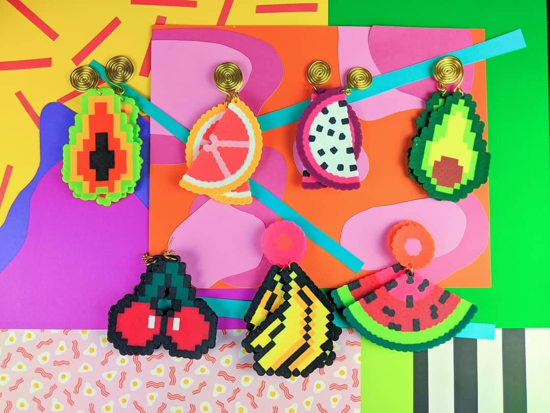 She's Behind BHAM's Most Colorful (and Secretly Nostalgic) Jewelry Line - 2 Colorful pixel art fruit keychains, crafted by a talented jewelry designer, with papaya, grapefruit, dragonfruit, avocado, cherries, banana, and watermelon on a vibrant patterned background.