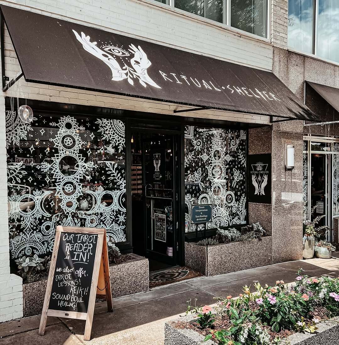 Storefront of Ritual + Shelter with black and white mystical designs on the windows, a sign advertising tarot readings by Jennifer Dorman, and plants lining the sidewalk in front.