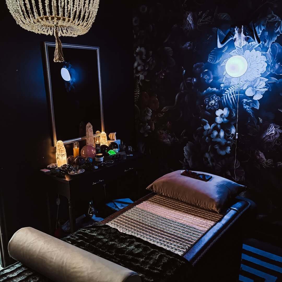 A dimly lit room by Ritual + Shelter with a massage table, a side table holding crystals and candles, a beaded chandelier, and floral-patterned dark walls softly illuminated by blue light.