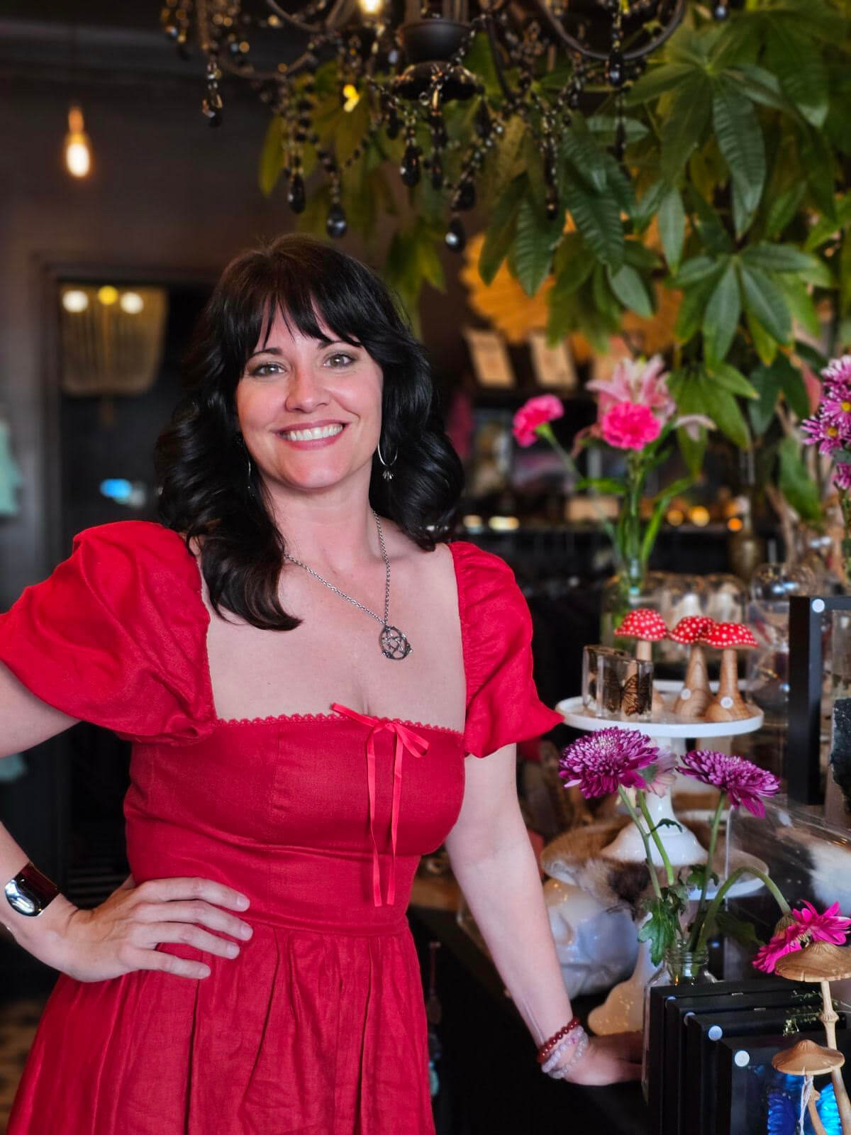 Jennifer Dorman stands in a red dress smiling with one hand on her hip. Surrounded by flowers and decorative items at her shop, Ritual + Shelter.