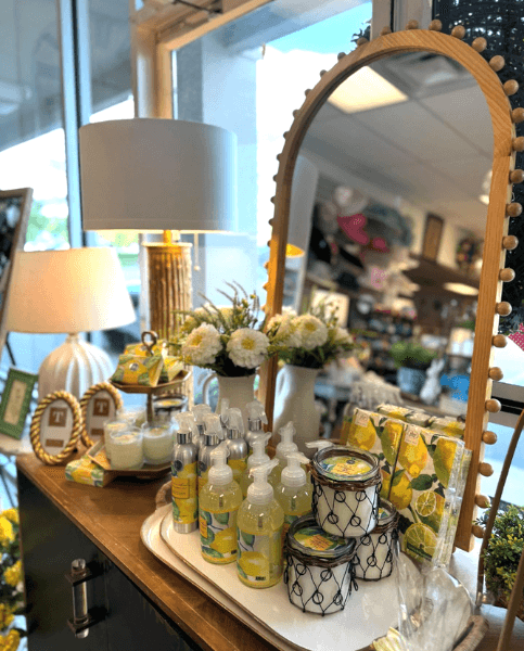 A wooden table in Louisville KY displays lemon-themed soaps and lotions, decorative jars, towels, and a vase of white flowers—perfect for boutiques—set before a mirror and two lamps near a window.