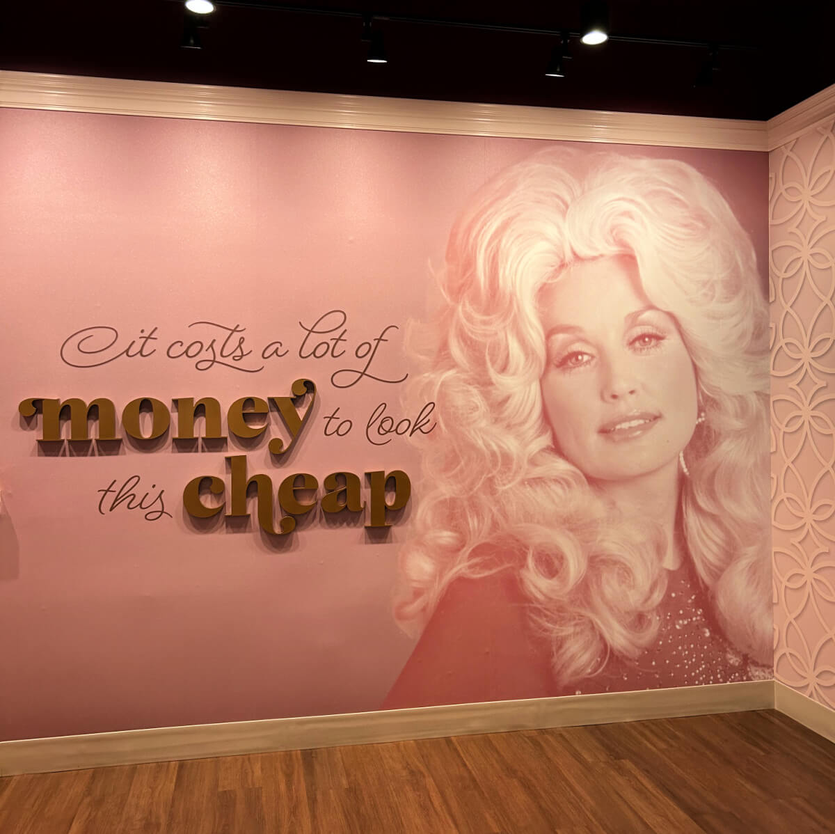 A pink wall at Dollywood features a portrait of Dolly Parton and the quote, “It costs a lot of money to look this cheap,” with part of the text in gold lettering—making it one of the top things to do for fans and selfie-seekers alike.