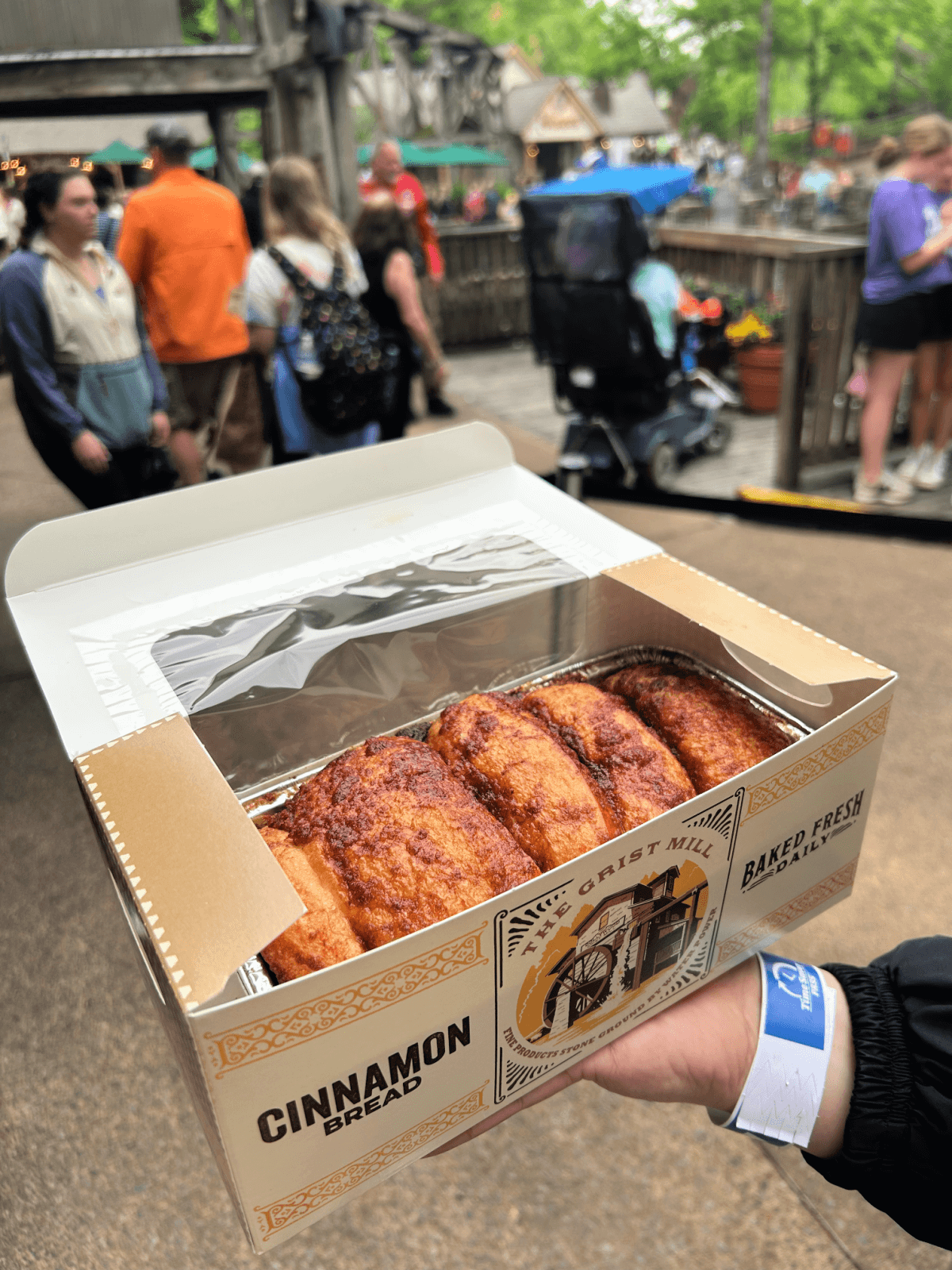 A hand holds an open box of cinnamon bread outdoors at Dollywood, with people walking and milling around in the background—a must-try treat among the many things to do at the park.