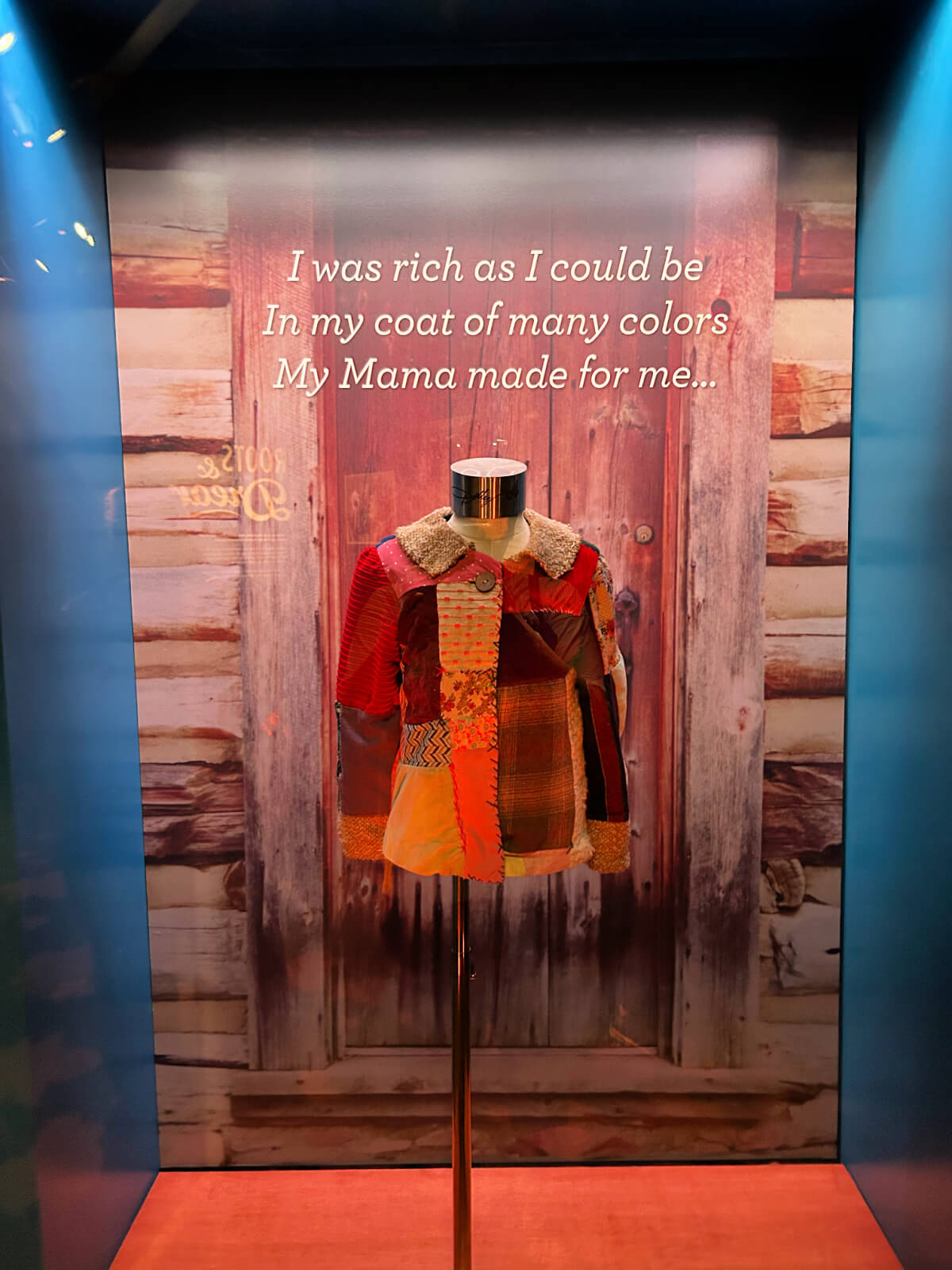 A colorful patchwork coat displayed on a mannequin at Dollywood, with a rustic wooden door backdrop and the quote, "I was rich as I could be in my coat of many colors My Mama made for me..."—a must-see among things to do.