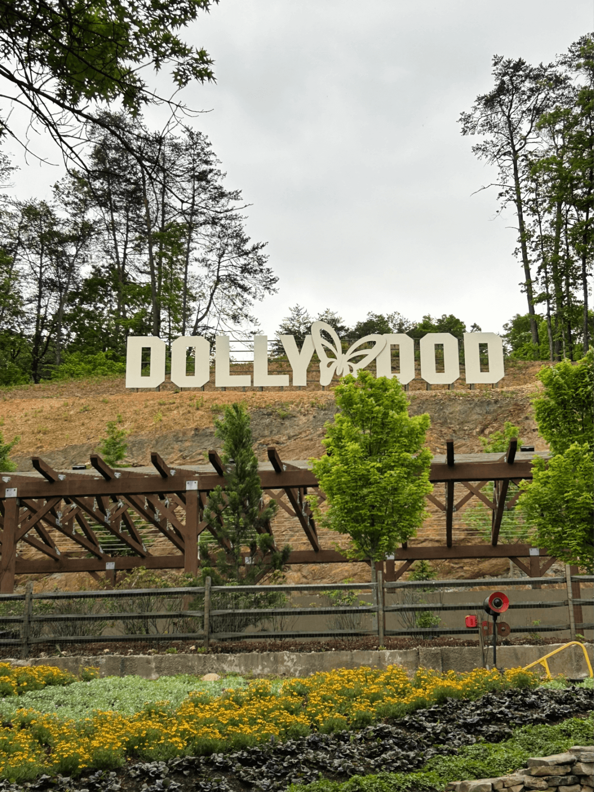 A large white “Dollywood” sign with a butterfly symbol stands on a hillside surrounded by trees and flowers under a cloudy sky, inviting visitors to explore the many things to do at Dollywood.