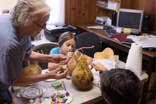 An adult and two children paint and decorate a large gourd at a table in a craft room, surrounded by art supplies and a computer—capturing the creative spirit of Hands-On Gatlinburg.