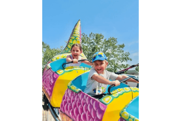 Two children ride a colorful dragon-themed amusement park ride outdoors on a sunny day in Corinth MS during the Slugburger Festival, both smiling and holding onto the handles.