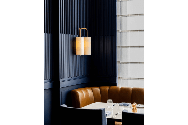 A Blue Aster restaurant booth with a brown leather banquette, white dining table, dark vertical paneled walls, and a modern wall sconce. Light filters through sheer blinds—perfect for a memorable Father’s Day Dinner.