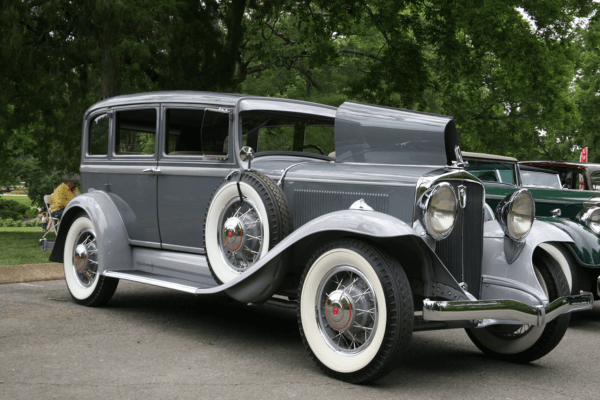A vintage gray car with whitewall tires is parked outdoors at Cheekwood, its hood partially open, surrounded by trees and other classic cars.
