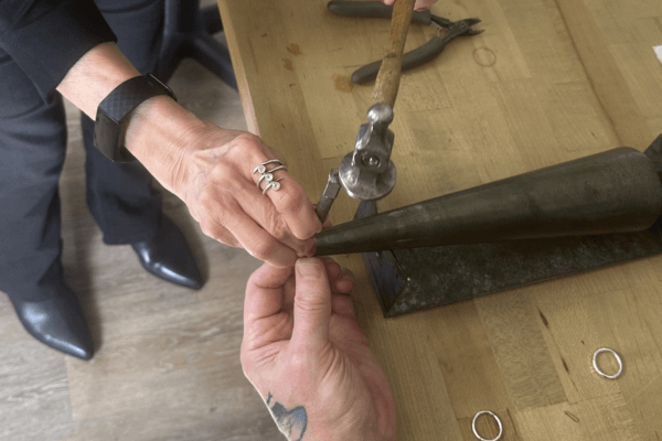 Two people work together to hammer a metal ring onto a mandrel on a wooden table, surrounded by tools and rings—capturing the creative spirit of Margaret Ellis Jewelry and their summer jewelry making classes.