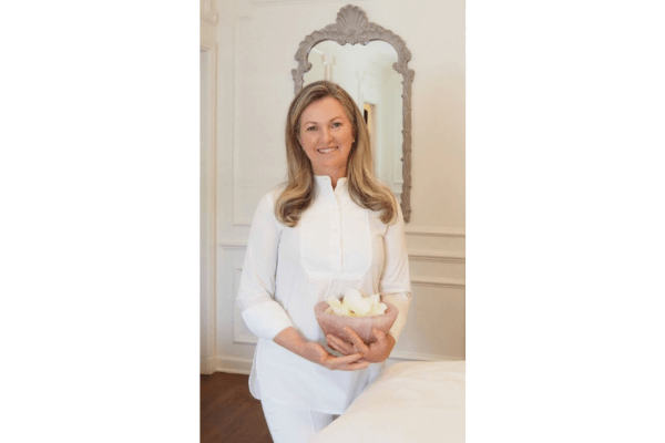 A woman in a white outfit stands indoors, holding a bowl of white flowers, with a decorative mirror on the wall behind her—perfectly capturing the serene essence of a Tammy Fender launch.