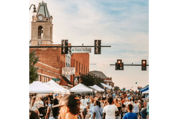 A large crowd walks among vendor tents at a street fair in a downtown area, with a clock tower and public parking sign visible in the background.