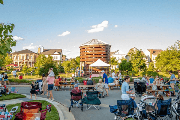 Residents gather outdoors in Stephens Valley for a lively community event, enjoying BBQ at picnic tables while children play and bluegrass music fills the air around a central structure.