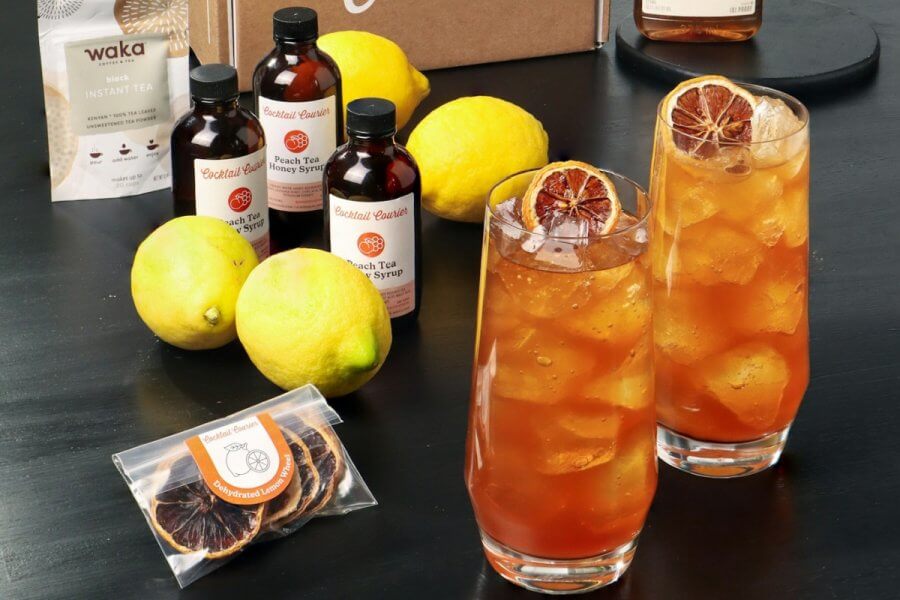 Two tall glasses of iced tea with dried citrus garnish sit on a table, surrounded by lemons, bottles of syrup, a coffee packet, and a small bag of dried orange slices—perfect for celebrating Father's Day in refreshing style.
