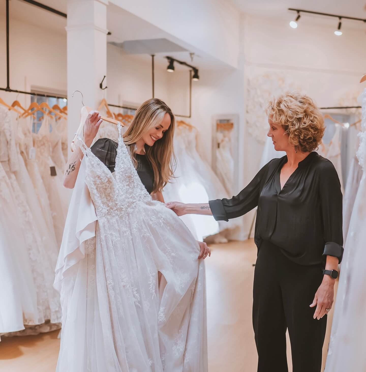 Two women stand in a Louisville, KY bridal shop, one holding up a white wedding dress while the other admires it. Rows of elegant gowns hang in the background of this charming boutique.