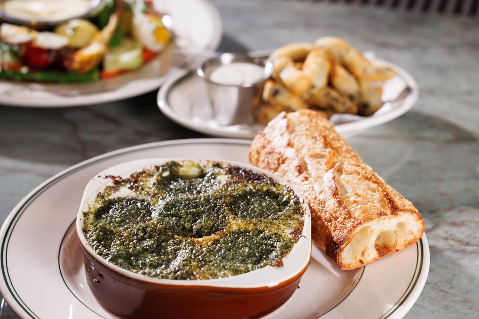A plate with baked escargot in garlic herb butter, a toasted baguette, and a side of fried pickles with dipping sauce in the background at one of the exciting new Louisville restaurants featuring a unique menu.