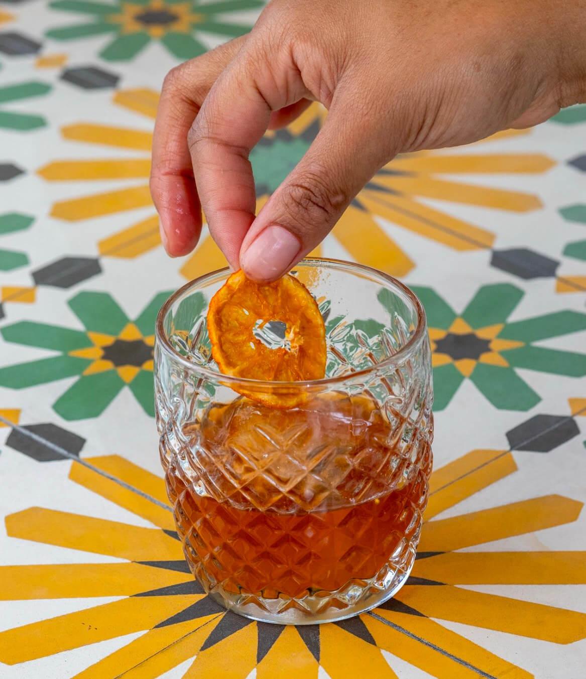 A hand places a dried orange slice into a glass of brown liquid on a patterned table with yellow and green geometric designs, showcasing one of the creative drinks from new menus at Louisville restaurants.