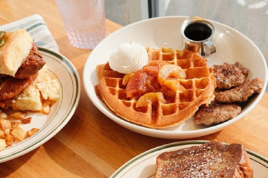 A plate with a waffle topped with whipped cream and fruit, syrup, and two sausage patties sits on a wooden table alongside other breakfast dishes—a tasty find at one of the best new restaurants in Louisville.