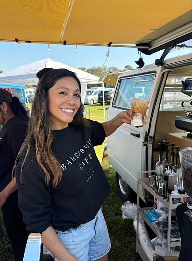 A woman standing outdoors by a van smiles and holds up an iced coffee drink at a cafe stall, with coffee supplies visible nearby.