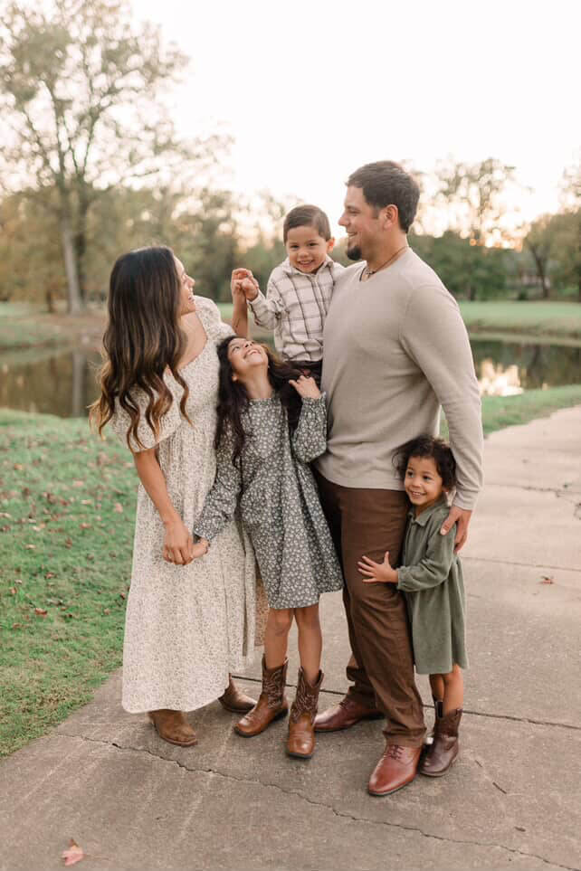 A family of five stands on a sidewalk in a park near West Tennessee, smiling and interacting with each other. There is a pond and trees in the background.