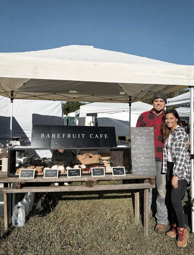 Two people stand in front of a stall at an outdoor market in West Tennessee, displaying baked goods and a chalkboard menu under a white canopy.