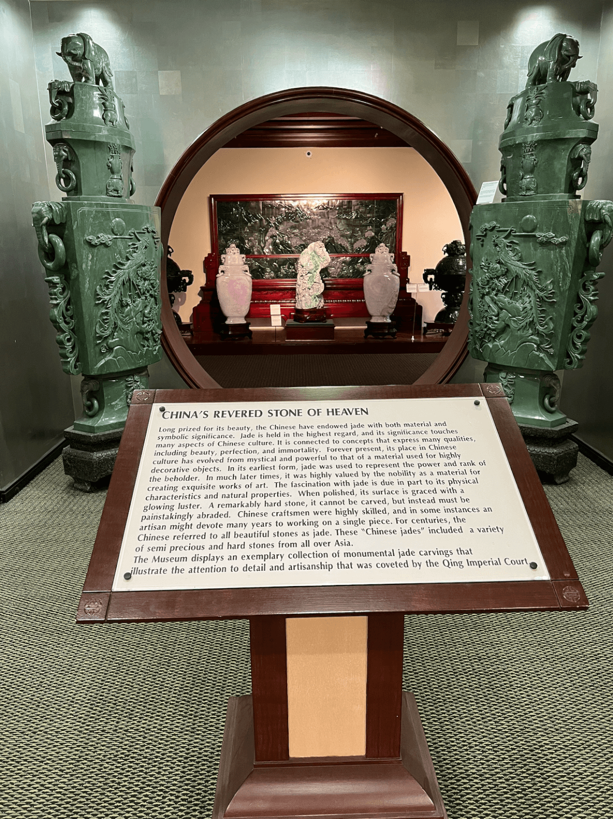 A museum display at the Belz Museum, features a sign titled "China's Revered Stone of Heaven" in front of jade sculptures and ornate green columns, with a framed artwork visible in the background.