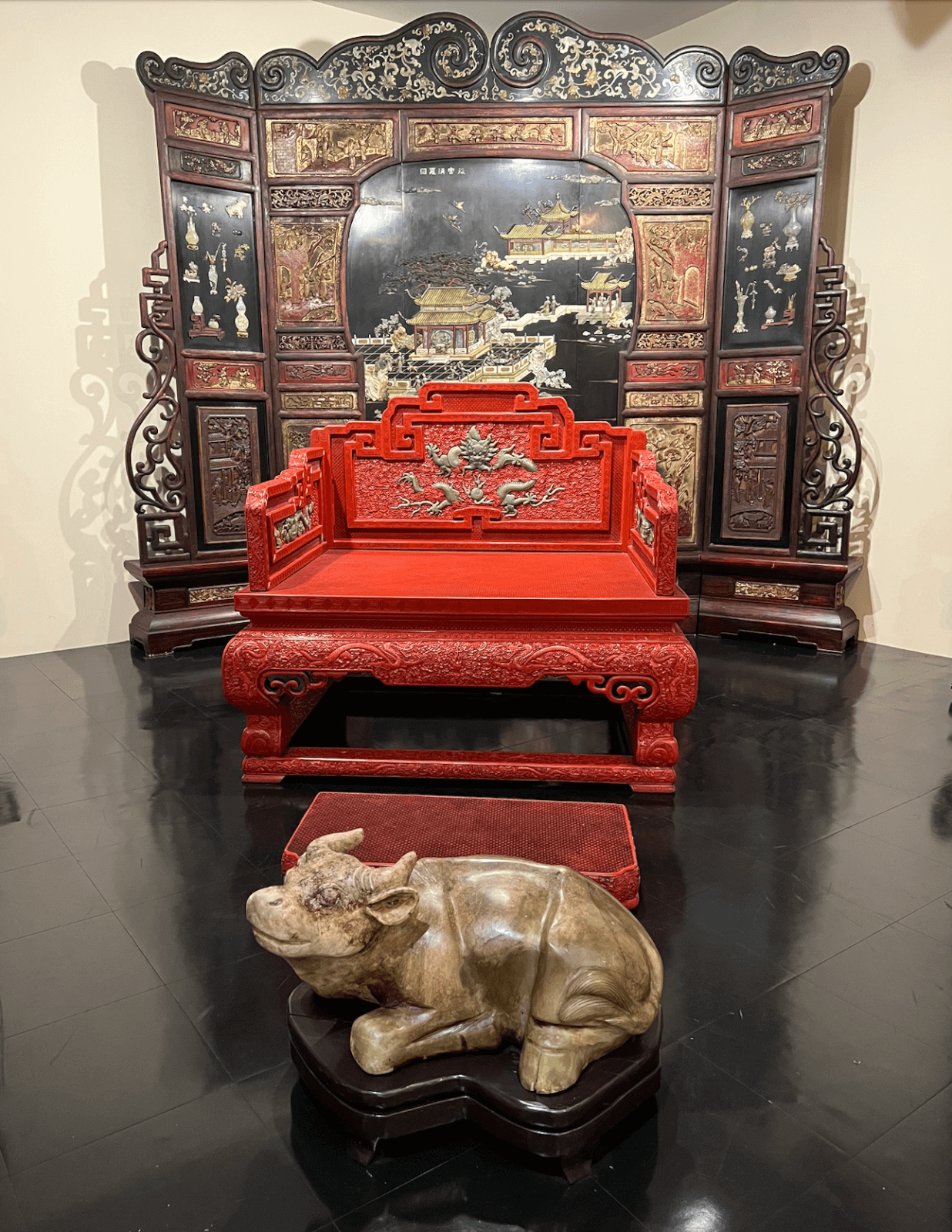 Ornate red wooden bench with dragon carvings stands before a decorative painted folding screen at Memphis's best-kept secret, the Belz Museum; a stone buffalo sculpture is positioned in the foreground on a dark floor.