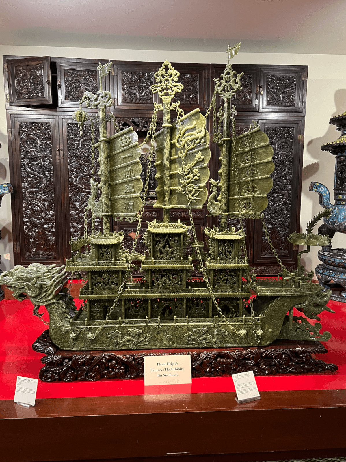 Ornate jade ship sculpture with detailed carvings and multiple sails on display at the Belz Museum, set on a dark wooden base with informational plaques in front.