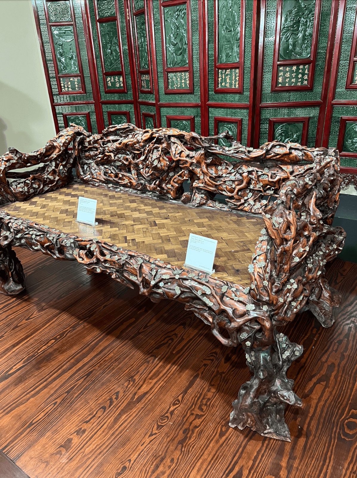 Ornate wooden bench with intricate carvings and a woven seat, displayed indoors on a wooden floor at the Belz Museum in Memphis—one of the city's best-kept secrets—with informational placards and a carved screen in the background.
