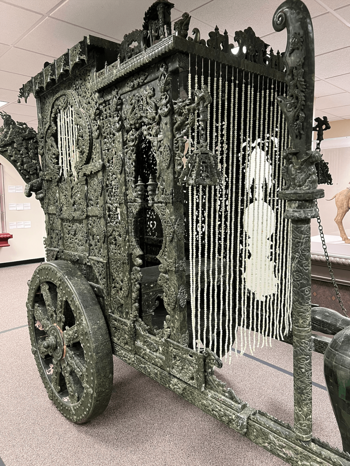 Intricately carved green stone chariot with beaded curtains, decorative details, and two large wheels, displayed indoors on a carpeted floor at the Belz Museum.